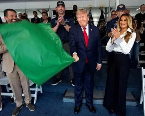 Donald Trump, wearing a suit, smiles as he waves a green flag while standing next to Melania Trump, who is clapping